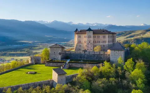 Castelli Val di Non, un imponente maniero su una collina verdeggiante, con montagne innevate sullo sfondo.