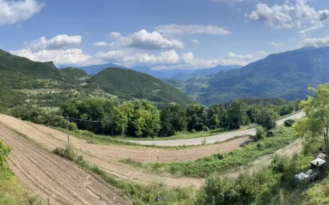Panoramica del passo di Santa Barbara a Trento, con campi coltivati, alberi verdi e montagne sullo sfondo.