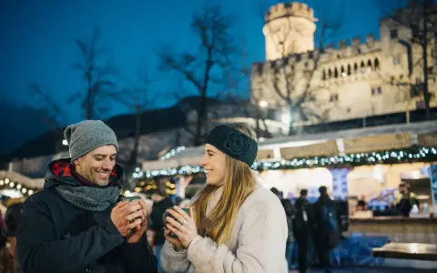 Coppia sorridente con tazze fumanti al mercatino di Natale di Trento inverno, con il castello illuminato sullo sfondo.
