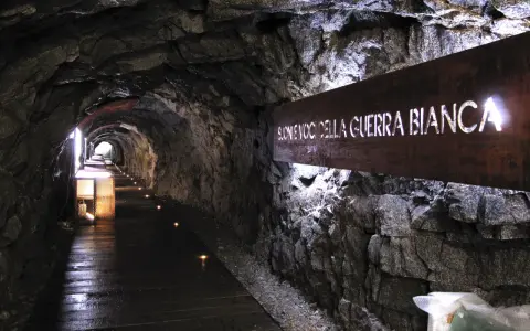 Un suggestivo tunnel roccioso illuminato, con un cartello che recita "Suoni voci della Guerra Bianca". Un percorso che sembra condurre alla galleria paradiso.