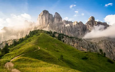 Sentiero serpeggiante su un prato verdeggiante ai piedi di un imponente gruppo montuoso tra Veneto e Trentino Alto Adige, con nuvole che accarezzano le cime rocciose.