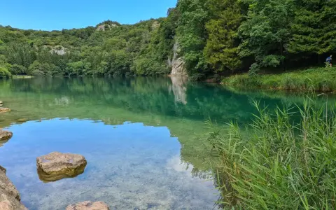 Acque cristalline del lago di Lavis riflettono la rigogliosa vegetazione. Rocce affiorano, un corridore si allena lungo la riva.