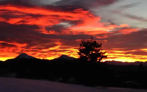 Un infuocato tramonto sulle Dolomiti, con nuvole rosse e arancioni che illuminano il cielo sopra le sagome scure delle montagne e di un pino solitario.
