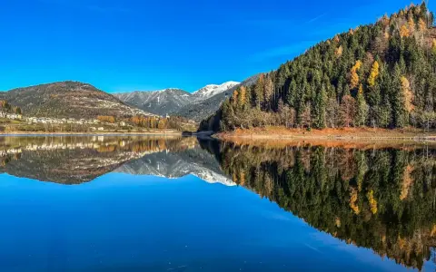 Il lago delle Piazze riflette montagne innevate, boschi autunnali e un villaggio sotto un cielo azzurro.