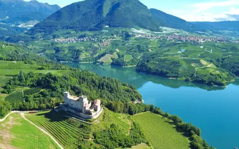Castello medievale su una collina verdeggiante, con vigneti a terrazze e un lago azzurro nel **Val di Non**.