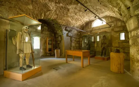 Interno di un museo in pietra con manichini in uniformi storiche, uno dei quali indossa un abito chiaro e porta un fucile, evocando un forte strino di battaglia.