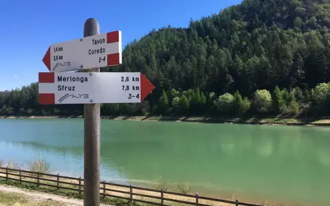 Cartello con indicazioni per Tavon di Coredo, Merlonga e Sfruz, con lago verde e bosco sullo sfondo.