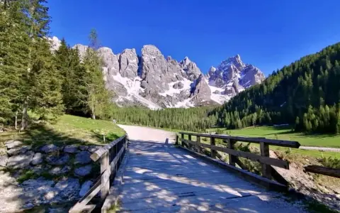 Ponte in legno su ruscello, sentiero per trekking Val Venegia, con montagne innevate e boschi verdi sotto un cielo azzurro.