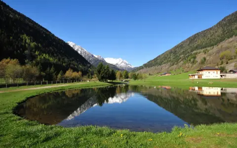 I laghetti di Vermiglio riflettono montagne innevate e un cielo azzurro. Un edificio moderno si specchia nell'acqua calma.