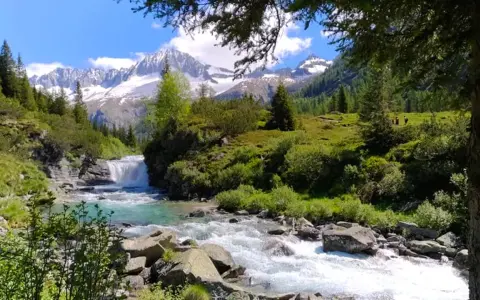Un torrente impetuoso scorre tra rocce e vegetazione lussureggiante nel **Val di Fumo**, con montagne innevate sullo sfondo.