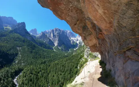 Sentieri panoramici vicino al Lago di Molveno, con un sentiero che costeggia una parete rocciosa e una vista mozzafiato sulle montagne e una cascata in lontananza.