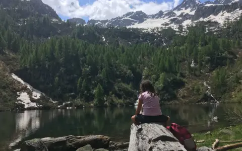 Bambina seduta su un tronco guarda il lago di Barco, circondato da montagne innevate e foreste di conifere.