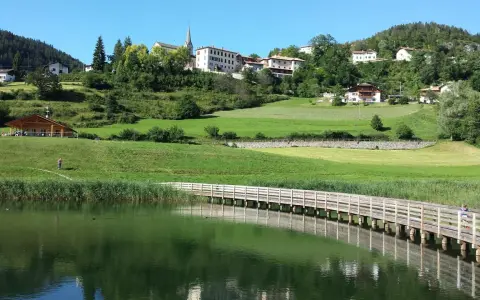 Una piacevole passeggiata dria al fos, con un ponte di legno che attraversa il lago e un villaggio sullo sfondo.
