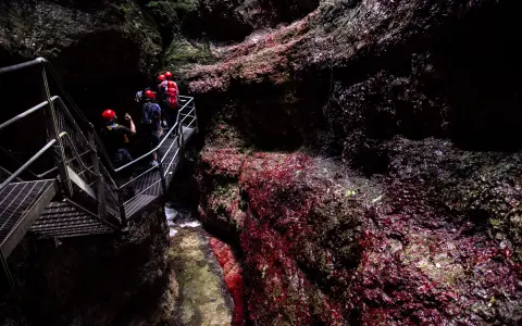 Esploratori con caschi rossi percorrono una passerella metallica nel **canyon Rio Sass fondo**, tra rocce scure e rosse.