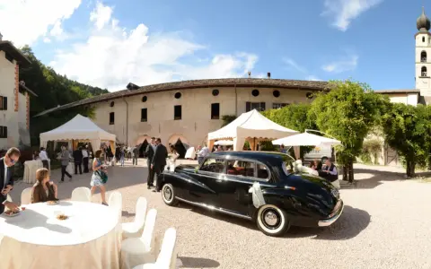 Matrimonio a Caldonazzo centro storico: auto d'epoca nera, tavoli allestiti, ospiti e un campanile sullo sfondo.