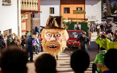 Gigantesca maschera pelosa con cappello marrone sfila al carnevale trentino. Un uomo con maschera grottesca trascina una catena.