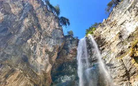 La cascata di Tret scende maestosa da una parete rocciosa imponente, sotto un cielo azzurro e limpido.