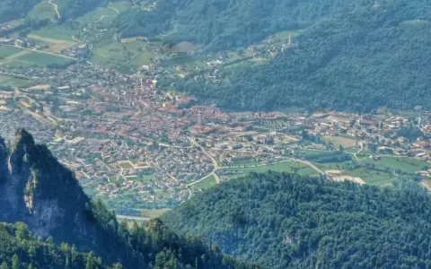 Vista aerea della Valsugana, Trento, con un paese incastonato tra montagne verdi e un picco roccioso in primo piano.