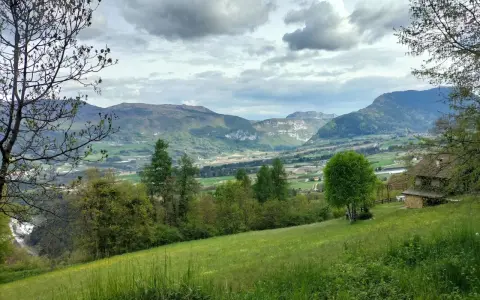 Prato verde e montagne sullo sfondo nella valle di Comano. Una casa con tetto in legno e un albero solitario.