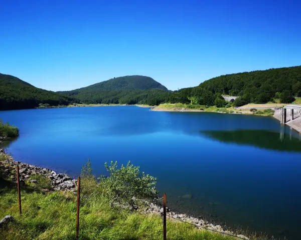 Vasto lago di Comano con acque blu intenso, circondato da colline verdi e una diga in cemento.