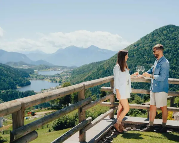 Coppia brinda con vino su un balcone con vista sulla **montagna di Pinè** e laghi.