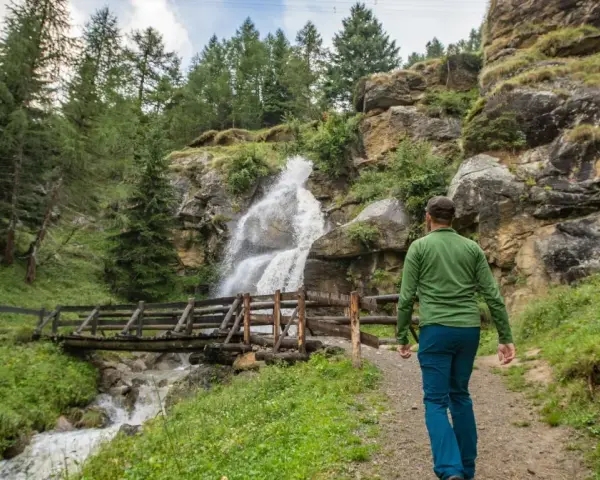 Un escursionista ammira una cascata nel Val di Sole in estate, con un ponte di legno che attraversa il torrente.