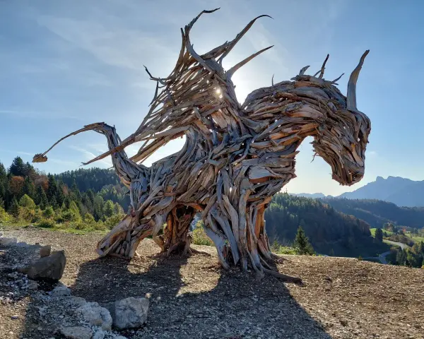 Un maestoso drago di Vaia, scolpito nel legno, domina un paesaggio montano con le ali spiegate verso il sole.