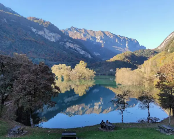 Il lago di Tenno riflette montagne imponenti e alberi autunnali. Due persone siedono su una panchina, godendosi la quiete.