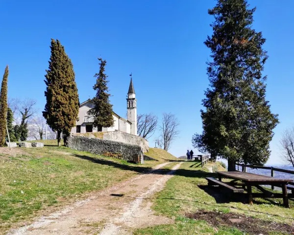 A church with a bell tower sits on a grassy hill, with tall trees and a picnic table in the foreground.