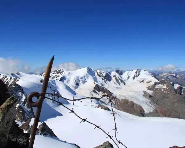 Vista mozzafiato dalla punta Linke, con cime innevate e un cielo azzurro intenso. Un vecchio piccone arrugginito e filo spinato segnano il confine.