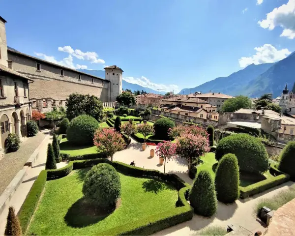 Giardini curati e fioriti incorniciano il maestoso Castel Buonconsiglio, con torri imponenti e montagne sullo sfondo.