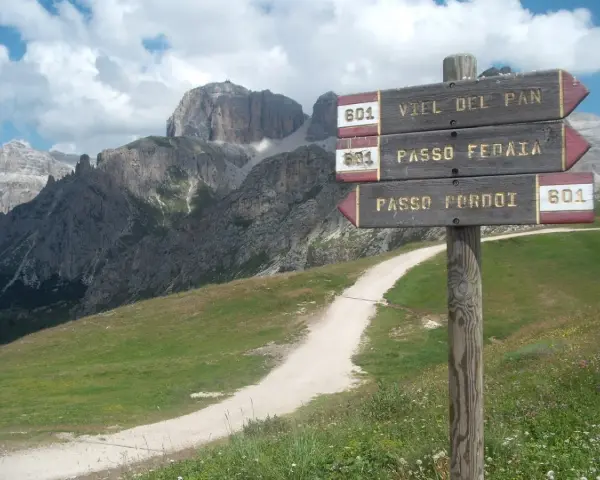 Cartello con indicazioni per Viel del Pan, Passo Fedaia e Passo Pordoi. Sentiero in salita tra prati verdi e montagne imponenti.