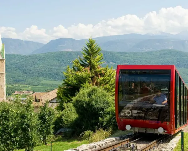 Funicolare rossa in Trentino Alto Adige, con campanile e montagne sullo sfondo. Un'ottima idea per scoprire cosa vedere!