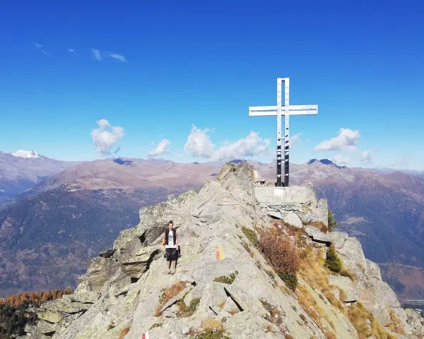 Escursionista raggiunge la vetta con la croce della pace, circondata da montagne maestose sotto un cielo azzurro.