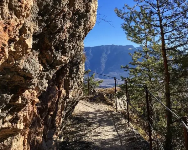 Sentiero margherita: un cammino roccioso con parapetto metallico, alberi e montagne sullo sfondo.