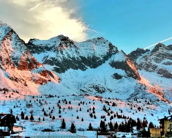 Montagne innevate al tramonto sul Passo del Tonale, con alberi sparsi e un hotel.