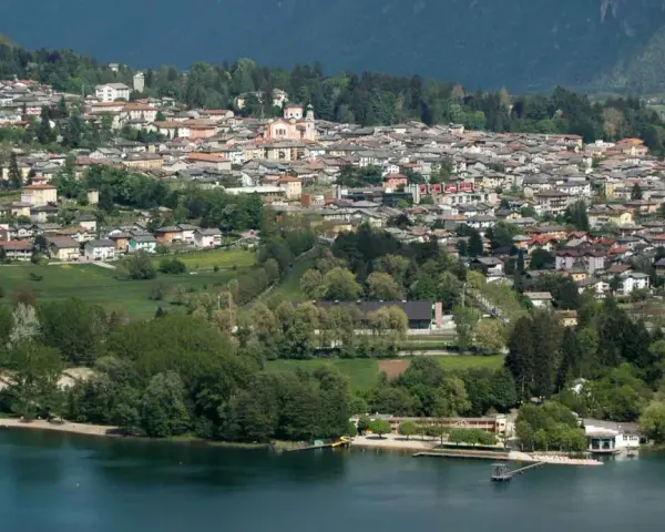 Vista panoramica del **lago di Levico** con il paese arroccato sulla collina. Alberi verdi e case colorate si specchiano nell'acqua tranquilla.