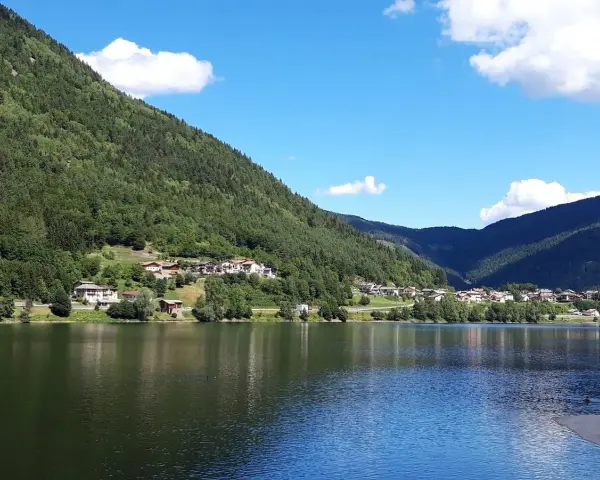 Il **lago di Serraia** riflette le montagne boscose e un villaggio alpino sotto un cielo azzurro con nuvole bianche.