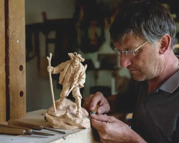 Maestro intaglia un pastore con bastone, un esempio di artigianato legno trentino.