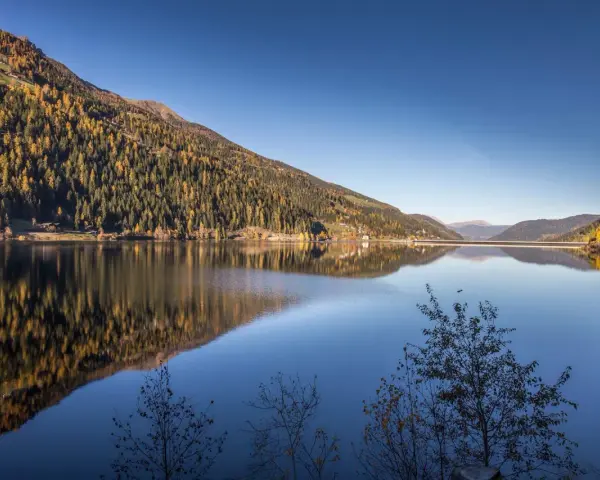 Autunno dorato sul **Val d'Ultimo lago**, con boschi che si specchiano nelle acque calme e un cielo azzurro intenso.