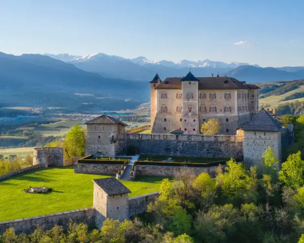 Castelli Val di Non, un imponente maniero su una collina verdeggiante, con montagne innevate sullo sfondo.