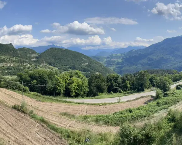 Panoramica del passo di Santa Barbara a Trento, con campi coltivati, alberi verdi e montagne sullo sfondo.