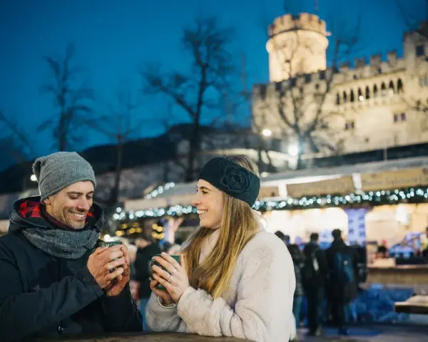 Coppia sorridente con tazze fumanti al mercatino di Natale di Trento inverno, con il castello illuminato sullo sfondo.