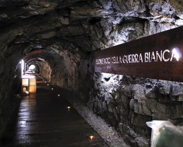 Un suggestivo tunnel roccioso illuminato, con un cartello che recita "Suoni voci della Guerra Bianca". Un percorso che sembra condurre alla galleria paradiso.