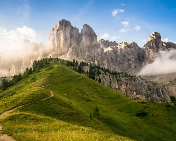 Sentiero serpeggiante su un prato verdeggiante ai piedi di un imponente gruppo montuoso tra Veneto e Trentino Alto Adige, con nuvole che accarezzano le cime rocciose.