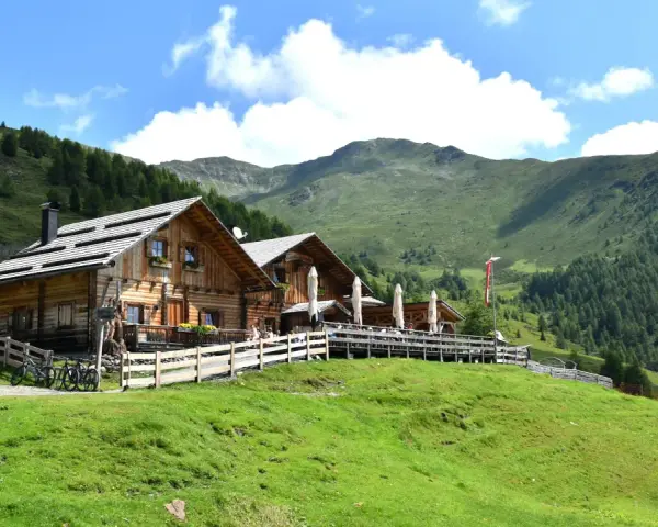 Rifugio in legno con tavoli all'aperto, ideale per un giro delle malghe in Val di Non. Montagne verdi e cielo azzurro.