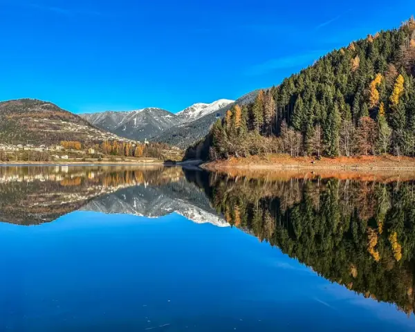 Il lago delle Piazze riflette montagne innevate, boschi autunnali e un villaggio sotto un cielo azzurro.