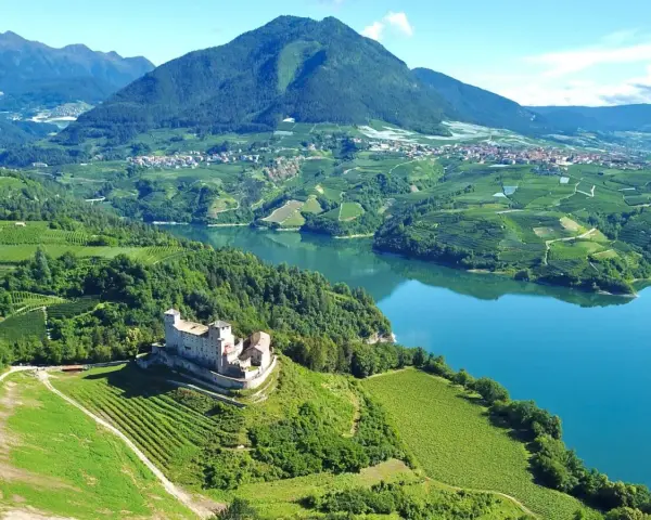 Castello medievale su una collina verdeggiante, con vigneti a terrazze e un lago azzurro nel **Val di Non**.