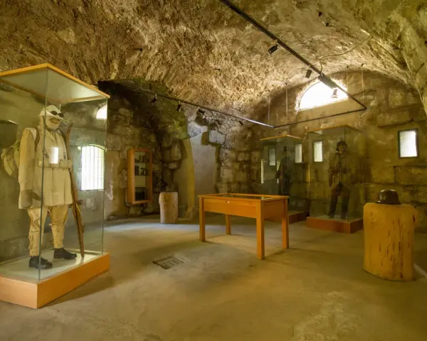Interno di un museo in pietra con manichini in uniformi storiche, uno dei quali indossa un abito chiaro e porta un fucile, evocando un forte strino di battaglia.