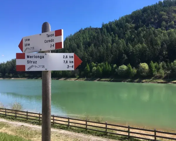 Cartello con indicazioni per Tavon di Coredo, Merlonga e Sfruz, con lago verde e bosco sullo sfondo.
